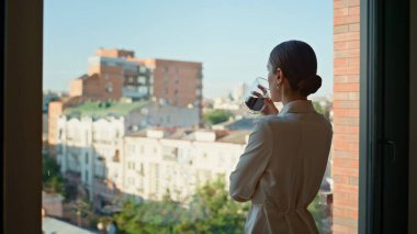 İş kadını kahvesini yudumluyor panoramik ofis penceresinden şehir manzarasına bakıyor. Şık bir iş kadını tatilde tek başına aromatik içecek içiyor. Rahatlamış kadın yönetici şehir manzarasının tadını çıkarıyor