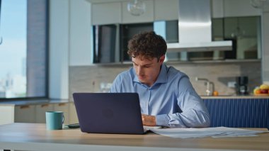 Mutfakta çalışan odaklanmış iş adamı dizüstü bilgisayarın yakın çekimini yapıyor. Modern ofis belgeleriyle ilgilenen ciddi bir adam. Yerel masadaki evrak işleri ve dijital görevler üzerinde yoğunlaşan uzaktan çalışan.