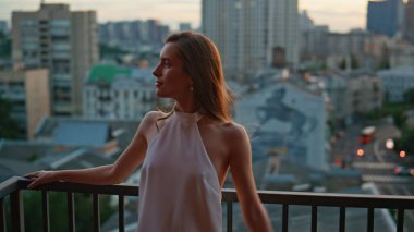 Esmer görünümlü şehir manzaralı çatı katı balkonuna yakın. Zarif beyaz elbiseli şehvetli bir kadın terasta tek başına dinlenirken korkuluklara yaslanıyor. Gülümseyen kadın şehrin alacakaranlığının tadını çıkarıyor özgürlüğünü hissediyor.