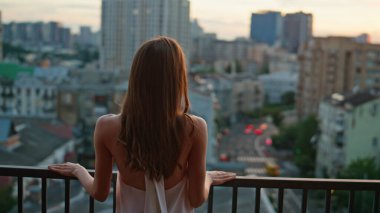 Alacakaranlık şehri manzarasında balkonda poz veren çekici bir model. Teras korkuluklarına yaslanmış zarif beyaz elbiseler giymiş rahat bir kadın. Gülümseyen esmer, çatıda durmuş, manzaraya bakıyor.