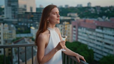 Çatıdaki balkonda duran muhteşem kadın elinde şarap kadehini tutuyor. Lüks elbiseli zarif bayan akşam manzarasına bakıyor. Güzel esmer, dingin anın tadını çıkarıyor. Terasta tek başına dinleniyor..