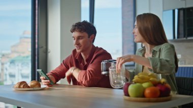 Gülümseyen eş, rahat kocasına kahve getiriyor. Parlak mutfakta akıllı telefon tutuyor. Mutlu çift birlikte oturup kahvaltıda cep telefonuna bakıyor. Cep telefonu fotoğrafı gösteren neşeli bir adam.