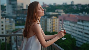 Zarif giyimli kadın balkonda şampanyayla sakin bir akşamın tadını çıkarıyor. Şık bir bayan şarap kadehi tutarak teras korkuluklarına yaslanıyor. Alacakaranlıkta harika bir esmer kamera..