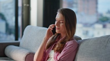 Ev kızı cep telefonuyla konuşuyor. Dairedeki kanepeye yakın oturuyor. Mutlu kadın sabah telefon konuşmasından zevk alıyor. Evde rahat ve rahat hissediyor. Neşeli bayan oturma odasını arıyor..