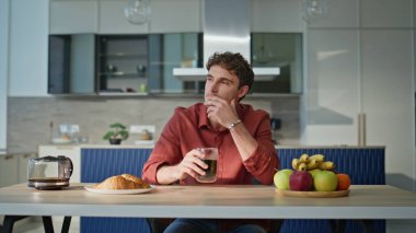 Kahve bardağını elinde tutan dalgın adam mutfak masasında tek başına oturuyor. Modern apartman mutfağında taze sabah içkisi içen düşünceli bir adam. Gevşek kahvaltı için taze kruvasan ve meyve.. 