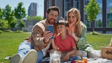 Piknikte cep telefonuyla konuşan neşeli bir aile. Çocuk sahibi mutlu çift, yeşil çimlerin üzerinde oturan modern bir aletle görüntülü görüşme yapıyorlar. Çocuklu neşeli ebeveynler akıllı telefona el sallıyor. 
