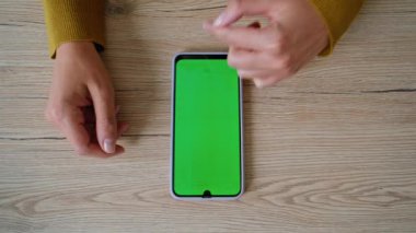 Ev masasında internet iletişimi için yeşil ekran akıllı telefonu kullanan kadın elleri. Ofiste kimliği belirsiz bir kadın cep telefonunu kaydırıyor. Krom tuşlu telefonda sosyal medyada gezinen kadın yönetici.