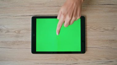 Yeşil ekranlı tableti işaret eden parmak işaretleri ahşap ev masasının üzerine yerleştirilmiş. Üst görüş alanı yakın. Meçhul bir kadın, internetteki uygulamayı tarıyor. İş kadını masadaki krom tuşlu dijital aygıta tıklıyor.