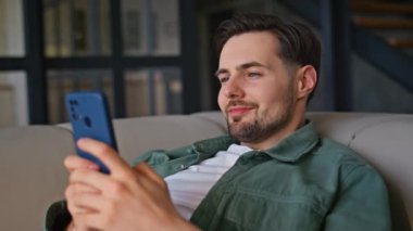 Yakın çekim yapan adam, akıllı telefondan hafif koltukta yalan söyleyen mesajlar atıyor. Hafta sonları sosyal medyada komik içerikler izleyen rahat ve mutlu bir adam. Oturma odasında cep telefonu kullanarak gülümseyen serbest çalışan.