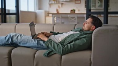 Dizüstü bilgisayarda çalışan bir adam koltukta rahat bir şekilde yatıyor. Bilgisayar işinden zevk alan serbest çalışan meşgul bir adam. Rahatlamış iş adamı bej oturma odasında e-posta yolluyor. Sakin yönetici dijital aygıtla yalnız takılıyor
