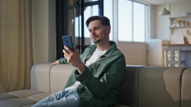 Evdeki adam yumuşak koltukta cep telefonu videosu izliyor. Modern bir dairede elinde akıllı telefon tutan yakışıklı bir genç. Gülümseyen müşteri koltukta cep telefonu uygulaması kullanarak online sipariş veriyor. 