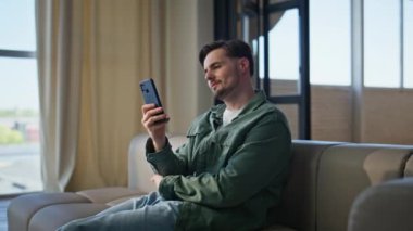 Hayalperest adam rahat koltukta akıllı telefondan mesaj okuyor. Gülümseyen yakışıklı adam cep telefonu ekranına bakıyor ve evde tek başına oturuyor. Kendine güvenen çekici kişi çevrimiçi tarama uygulamasını dairede satın alıyor 