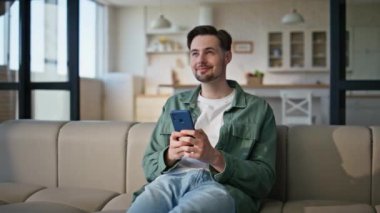 Gülümseyen adam cep telefonuyla samimi kanepeye mesaj yolluyor. Mutlu genç adam cep telefonu ekranına bakıyor ve ev oturma odasında SMS yaratıyor. Hayalperest erkek arkadaş akıllı telefondan kanepeye aşk mesajı atıyor.