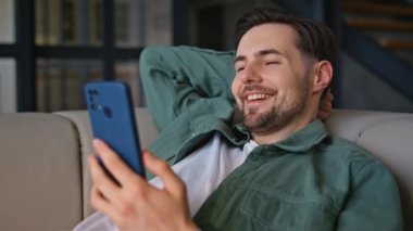 Rahatlatıcı cep telefonu görünümlü bir adam daire kanepesinde yatıyor, yakın plan. Gülümseyen mutlu adam akıllı telefondan komik internet içeriğini izlerken evde dinleniyor. Cep telefonu tutan rahat gülen işadamı 