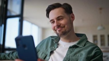 Gülen adam cep telefonuyla ev kanepesinde tek başına oturmaktan zevk alıyor. Akıllı telefon internet bağlantısıyla arkadaşlarını arayan yakışıklı adam videosu. Çevrimiçi toplantıda gülümseyen hipster selamlaması
