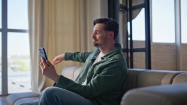 Gülümseyen hipster, evdeki kanepenin üzerinde cep telefonuyla video görüşmesini selamlıyor. Mutlu yakışıklı adam internette cep telefonu görüntülü web kamerasıyla arkadaşça buluşmanın tadını çıkarıyor. Akıllı telefon sanal sohbetini yalnız yapan olumlu adam.
