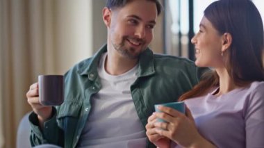 Kanepede elinde lezzetli kahve bardaklarıyla mutlu çift sohbeti. Neşeli genç eşler evdeki oturma odasında keyifli iletişimin tadını çıkarırlar. Gülümseyen çift aile tatilinde rahatlıyor.