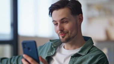 Modern oturma odasında akıllı telefondan canlı yayın yapan mutlu blogcu. İçerideki abonelere gülen neşeli bir adam. Koltuktaki cep telefonundaki video içeriğini kaydeden olumlu adam etkileyicisi