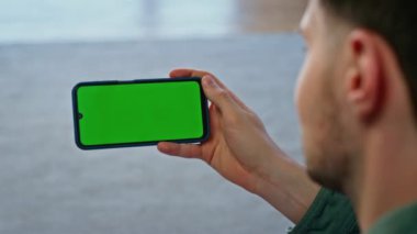 Oturma odasında akıllı telefonları tutan erkek elleri, yakın plan. Yeşil ekranda cep telefonuna bakıp sosyal medyayı izleyen tanınmamış bir adam. Dairede krom anahtar cep telefonu kullanan bir erkek. 