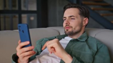Dondurucu esmer görünümlü cep telefonu bej kanepede uzanıyor. Rahat adam oturma odasında sosyal medya okuyarak kanepede dinleniyor. İçeride dinlenen mutlu gülen adam. Teknoloji ve insanlar kavramı