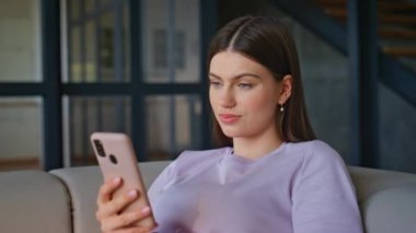Modern apartmandaki cep telefonundan mesaj okuyan ev kızı. Akıllı telefondan sosyal medyada gezinen gülümseyen genç bir kadın. İnternetten alışveriş yapan neşeli bir bayan oturma odasındaki koltukta cep telefonu kullanıyor. 
