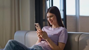 Neşeli kız cep telefonuyla sosyal medyada geziniyor. Ev kanepesinde dinleniyor. Akıllı telefondan video izleyip rahat koltukta oturan gülümseyen kadın. Neşeli ergen mesajlaşması cep telefonu sevinç verici.