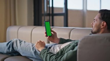Rahat adam rahat koltukta yatarken cep telefonu kullanıyor. Yakın plan. Sakallı adam, akıllı telefon yeşil ekranında yalnız başına fotoğraf izliyor. Günlük erkek sosyal medya hücresine göz atıyor