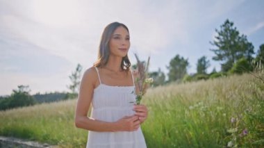 Kırsal model elinde çiçeklerle çayırda geziniyor. Yazın kır çiçeklerini taşıyan mutlu, gülümseyen bir kadın portresi. Mavi gökyüzü arka planında buketin keyfini çıkaran romantik bir kadın.