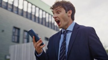 Heyecanlı kazanan modern sokak kapanışında cep telefonuna bakıyor. Akıllı telefon mesajı okuyan aşırı mutlu iş adamı başarıya duygusal tepki veriyor. Mutlu girişimci dans ediyor, eğleniyor ve eğleniyor..