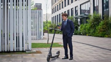 Modern kentsel alanda elektrikli scooter kullanan bir iş adamı. Şık takım elbiseli girişimci cep telefonu uygulaması kullanarak e-scooter 'a yaklaşıyor. Çevresel taşımada kendine güvenen ofis yöneticisi.