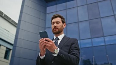 Gülümseyen yönetici akıllı telefonuyla ön cam binada dikiliyor. Zengin iş adamı modern kentsel alanda telefon güncellemelerini kontrol ediyor. Şehir caddesinde cep telefonu mesajını okuyan zarif bir yönetici..