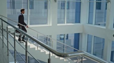 Motivasyonlu yönetici aşağıda modern güneşli ofis salonunda yürüyor. Sakallı işadamı elinde siyah bir çantayla merdivenlerden iniyor. Basarili bir CEO, merdivenlerden inerken el gözetimi yapiyor.