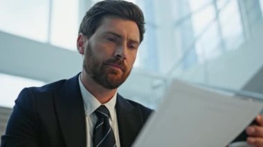 İş adamı, elinde kahve fincanıyla ofis merdivenlerinde oturan belgeleri inceliyor. Ciddi sakallı yönetici okuma sözleşmesi detaylarını yudumluyor. Evrakları inceleyen profesyonel bir adam.