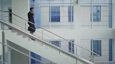 Profesyonel biri merdivenlerden inip cep telefonuyla konuşuyor. Başarılı iş adamı akıllı telefonu arıyor ve modern ofis binasına iniyor. Meşgul lider kol saatini kontrol ediyor. Merdivenlerden çantasıyla iniyor..