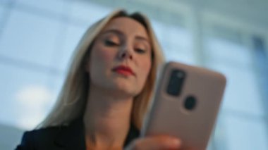 Sarışın iş kadını aydınlık ofis binasına yakın çekim yapan akıllı telefona odaklanmış. Modern şirket ortamında cep telefonu mesajlarına bakan profesyonel bir kadın yönetici. Çekici bayan patron cep telefonu gibi görünüyor..