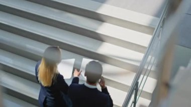 İş ortakları merdivenlerden modern ofis binasına çıkan belgeleri tartışıyor. İş kadınının evraklarını incelemek için iş adamını arıyorum. Şirket meslektaşları merdivenleri tırmanıyor ve konuşuyor..