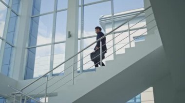 İş adamı aceleyle toplanıyor merdivenlerden iniyor modern ofis ortamında. Siyah evrak çantası taşıyan başarılı girişimci aşağıya iniyor. İşletme müdürü merdivenlerde nöbet tutuyor..