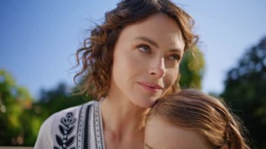 Şefkatli anne, güneşli çimenlikte kızının dinlenme parkını kucaklıyor. Gülümseyen esmer kadın, yeşillik içinde oturan anne dışarıda çocuklarla bağ kuruyor. Güzel, mutlu bir aile yaz çimenlerinde dinleniyor.