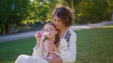 Yeşil güneşli parkta sabun renkli baloncuklar çıkaran ebeveyn çocuk portresi. Gülümseyen kıvırcık anne, güzel kızıyla oynuyor ve sıcak sonbahar gününün tadını çıkarıyor. Ebeveynlik duygusal bağlılık kavramı