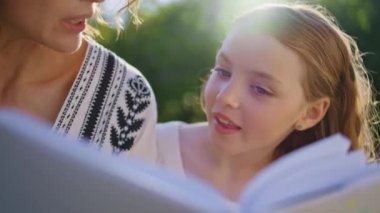 Anne çocuk kitap okuyor. Oturma alışkanlığı. Yakın çekim. Sevimli küçük kız haftasonunu yaz parkında geçiren anne babasıyla konuşuyor. Gülümseyen anne kız birlikte dinleniyor. Mutlu aile ilişkileri konsepti