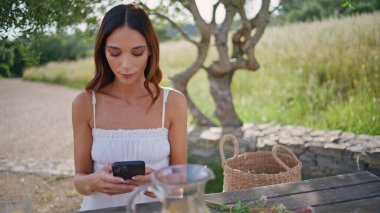 Kırsal kesimdeki bir kadın akıllı telefon ekranında pikniğe yakın oturuyor. Güzel kız cep telefonu uygulamasını yalnız başına kullanıyor. Romantik bayan sakin bir yerde hafta sonunun tadını çıkarıyor. Doğa konsepti 