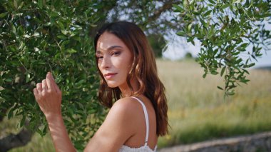 Yeşil bahçede gülümseyen, ışıl ışıl bir kadın. Doğanın yakın çekimlerinin keyfini çıkar. Zeytin ağacının yanında mutlu duran güzel bir kızın portresi. Beyaz elbiseli, kaygısız, şehvetli kadın gür çalıların önünde poz veriyor.