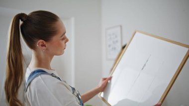 Parlak odada çerçeveli resmi yakından inceleyen bir kadın. Sevgi dolu çift modern apartmanı moda sanat eserleriyle dekore ediyorlar. Duvara resim asan genç eşler çağdaş alan yaratıyor.