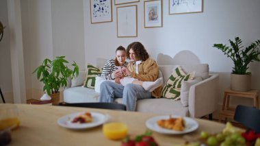 Çift kişilik cep telefonları rahat bir kanepede dinleniyor. Gülümseyen genç eşler akıllı telefondan mesaj okuyarak aile hafta sonunun keyfini çıkarıyorlar. Mutlu romantik çift telefon sosyal medyasında geziniyor..