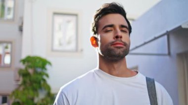Beyaz tişörtlü, güneşli bir sokakta duran kendine güvenen bir adam. Yakışıklı İspanyol adamın portresi. Kameraya güvenle dışarıda poz veriyor. Tıraşsız genç erkek ciddi ciddi kentsel çevreye bakıyor.. 