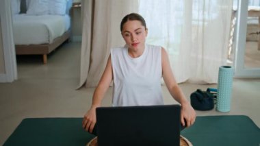 Sporcu kadın laptopta video taraması yapıyor. Modern ev içi yakın plan yoga minderinde oturuyor. Sıska bir kadın vücut ısıtması yapıyor ve bilgisayar eğitimi alıyor. Çekici bayan pilates egzersizi yapıyor. Monitöre bakıyor.