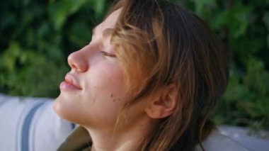 Huzurlu kadın yeşil bahçede gözlerini kapayarak rahatlama anının tadını çıkarıyor. Verandadaki kanepede oturan güzel bayan portresi. Genç esmer dinleniyor. Etrafı güneş ışığı ve yeşilliklerle çevrili..