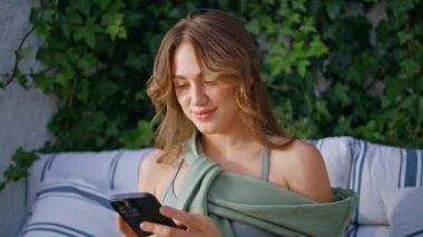 Cep telefonundan sosyal medyayı izleyen bir kız rahat bir terasa yakın oturuyor. Güneşli açık terasta cep telefonu mesajı okuyan gülümseyen bir kadın. Mutlu bayan internette sohbet etmeyi rahat koltukta dinlenmeyi sever.