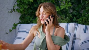 Kız rahat terasta taze portakal suyu içerek cep telefonundan zevk alıyor. Portre mutlu kadın konuşan akıllı telefon oturma terası çevresi yeşillikle çevrili. Güzel bayan telefonda konuşuyor, içkisini yudumluyor.