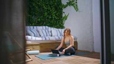 Verandada dizüstü bilgisayarla yoga yapan genç bir kadın. Açık havada spor paspası üzerine bilgisayar eğitimi alan aktif bir kız. Eğitimsel video izleyen esnek sporcu kadın kollarını uzatarak sağlığın tadını çıkarıyor.